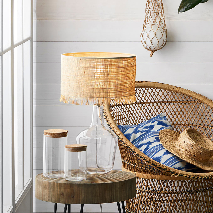 Lampe Ibiza en verre et raphia naturel placée sur une table en bois, avec fauteuil en osier et décor de style bohème. Dakar, Sénégal.